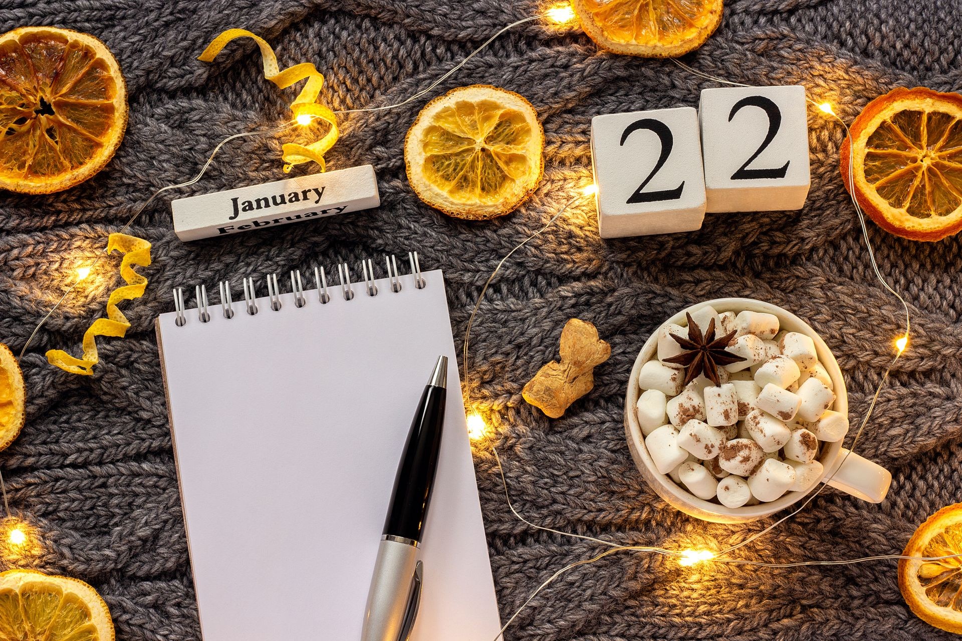 Winter composition. Wooden calendar January 22nd Cup of cocoa with marshmallow, empty open notepad with pen, dried oranges, light garland on grey knitted background. Top view Flat lay Mockup